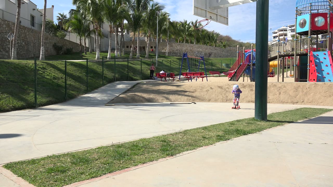 In Los Cabos, Mexico, a helmeted child rides along the right edge of a paved path while a red playground, palm trees, grass, and white buildings fill the background.