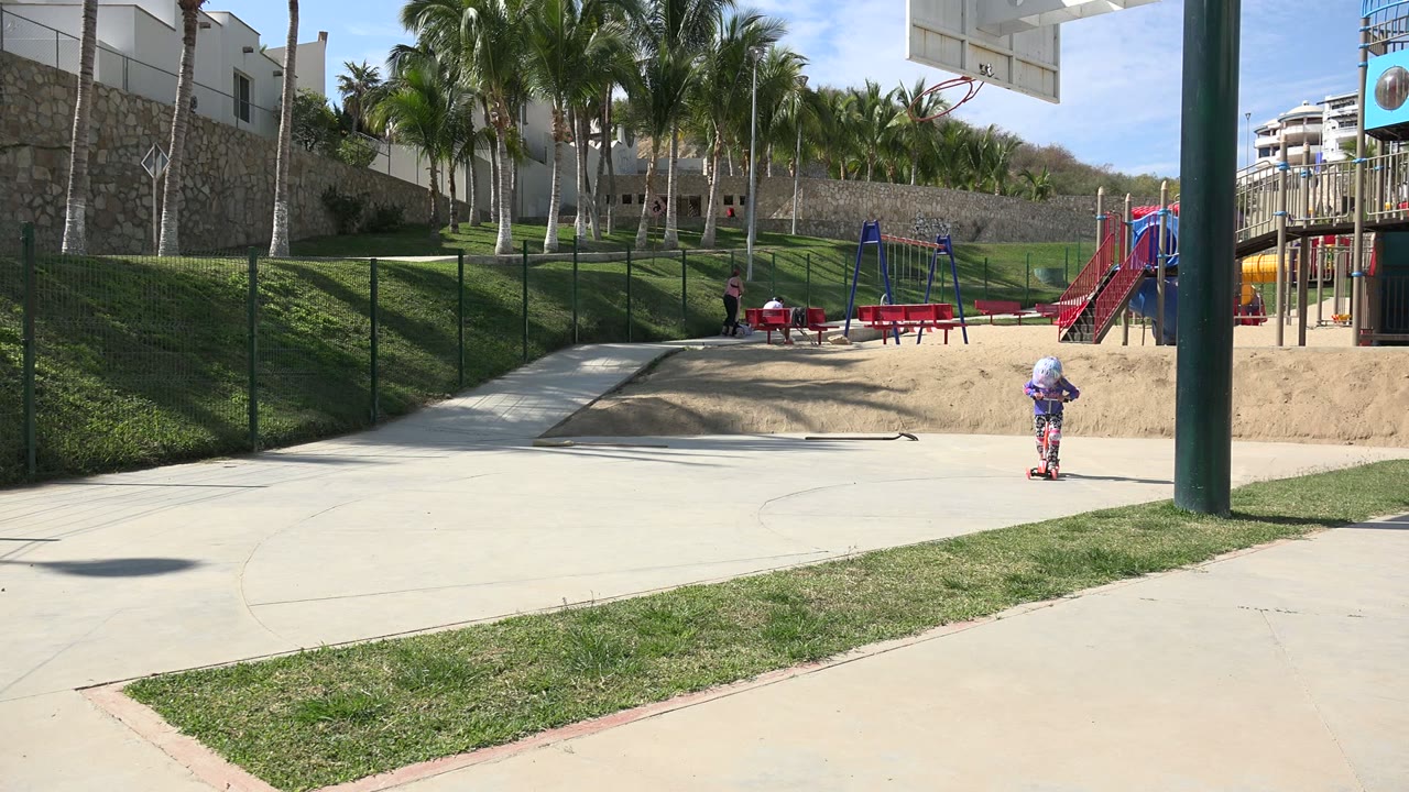 Wide park view with a small child riding near the right edge and playground behind.