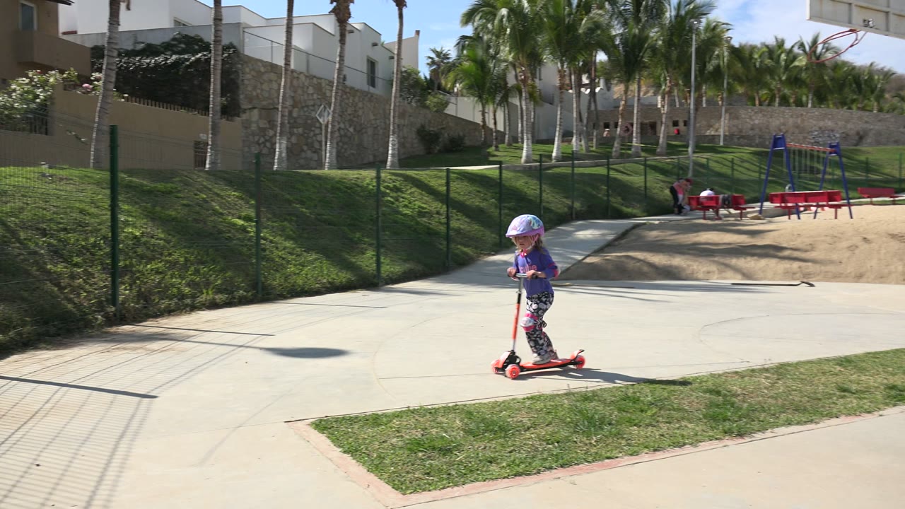 Small girl in a helmet rides a red scooter on a sunny park path.