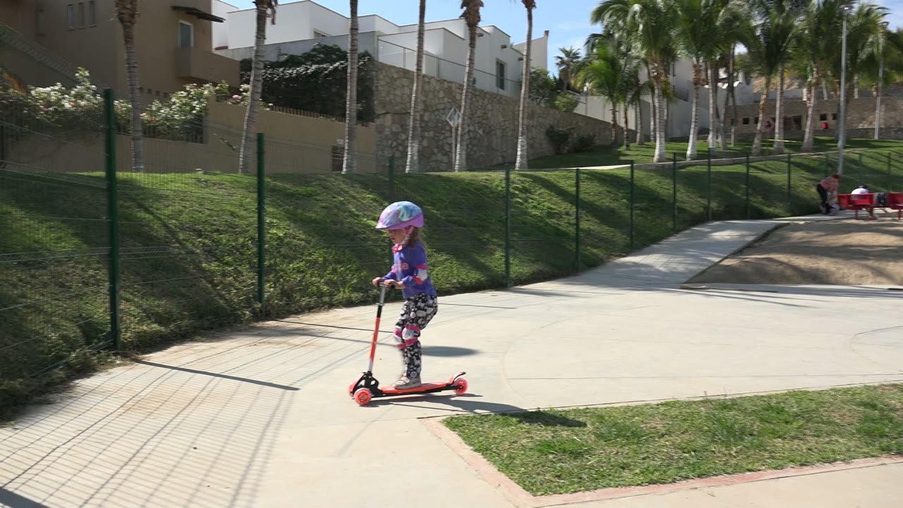 Helmeted girl rides a red scooter farther along the paved path in Los Cabos, Mexico, with a green fence, trimmed grass, palms, and pale buildings in the background.