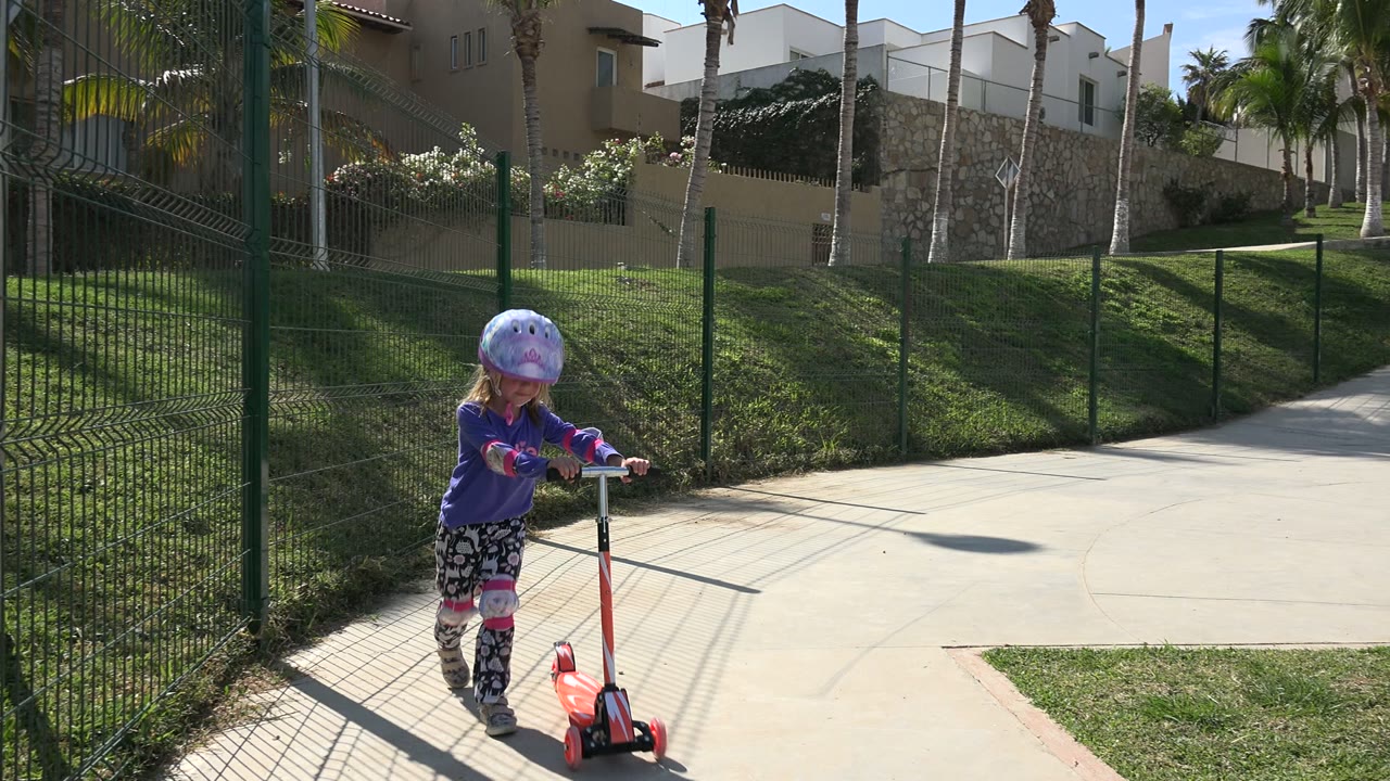 A helmeted child stands with a red scooter on a paved path beside a green fence in a sunny Los Cabos playground.
