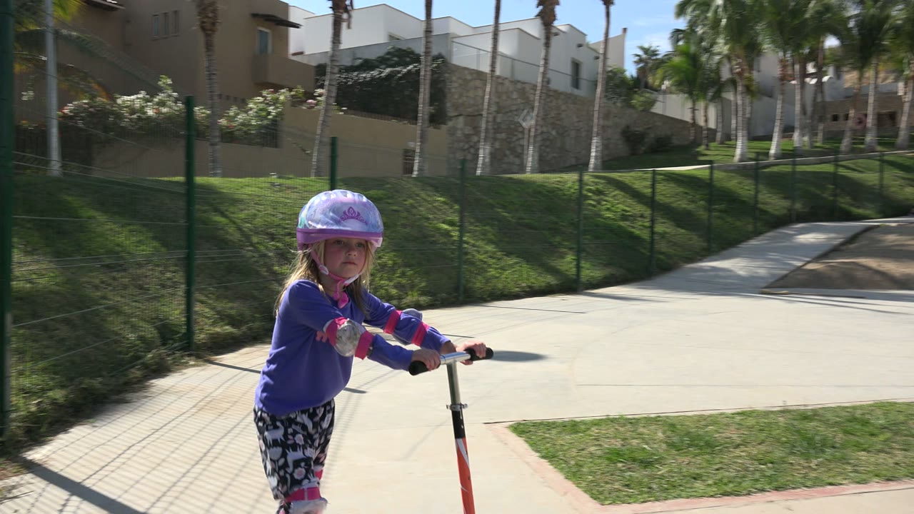 The child on the red scooter is closer to camera on the paved path, with green fencing, lawn, palms, and buildings behind.