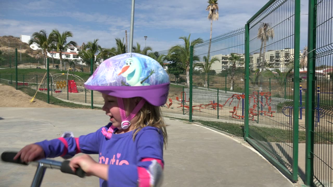 A young girl in a purple helmet is close to the camera with scooter handlebars at a fenced playground in Los Cabos, Mexico, with palms and play equipment behind her.