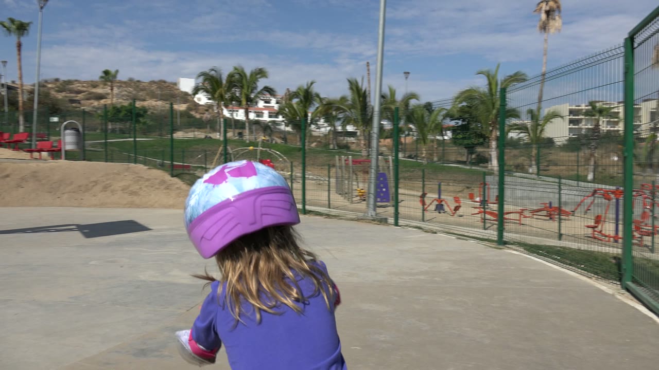 The child turns away from the camera in Los Cabos, Mexico, with her purple helmet and blue shirt prominent against green fencing, palms, and red playground equipment.