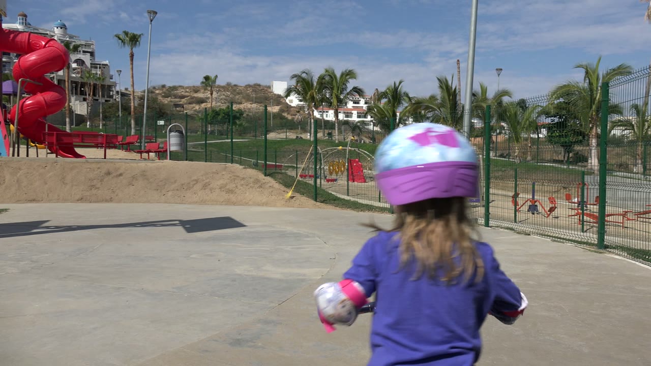 Girl moving away on a scooter near a fenced playground and red slide.