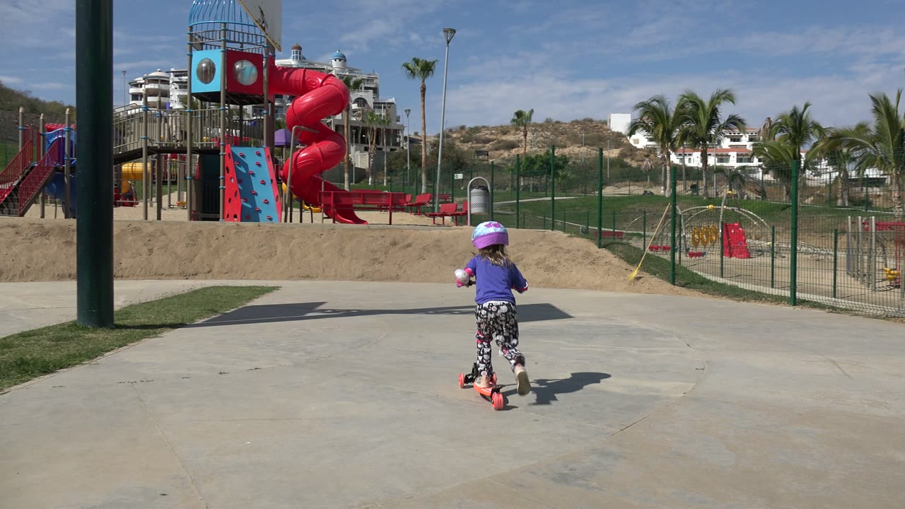 At 0:00 in Los Cabos, Mexico, a young girl in a purple shirt and helmet rides a red scooter away from the camera on open pavement toward a red playground slide.