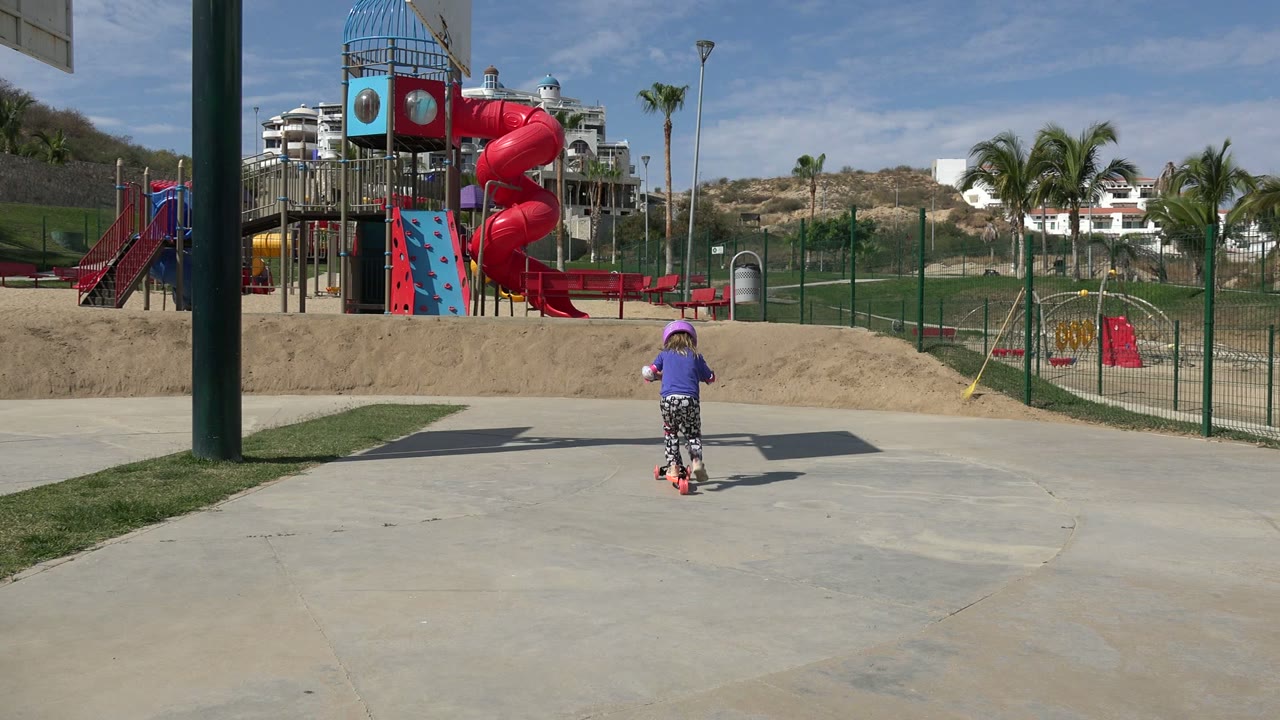 At 0:01 in Los Cabos, Mexico, the helmeted child rides farther into the paved area, with the red slide, fenced playground, palms, and hillside behind her.