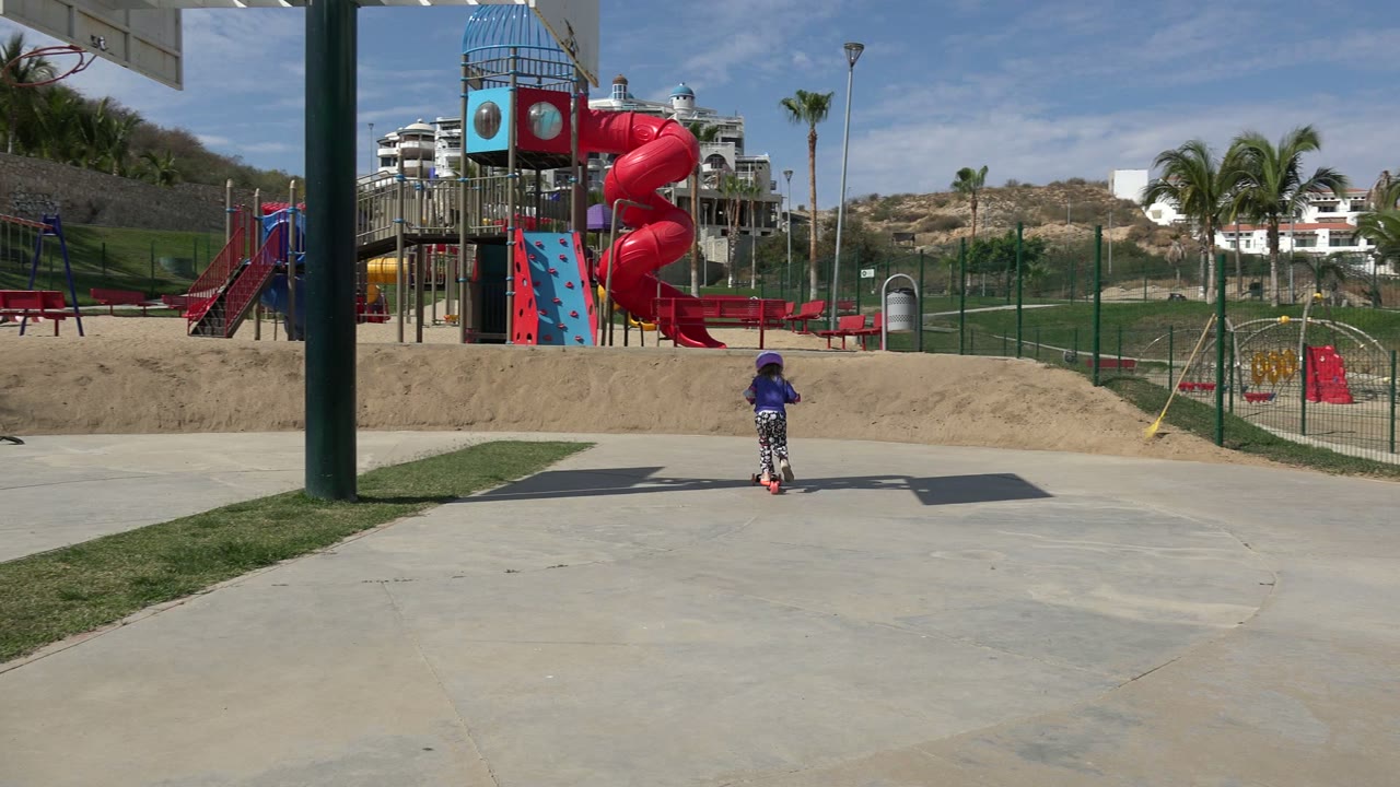 Small child rides a scooter deeper into a sunny park with playground equipment behind.