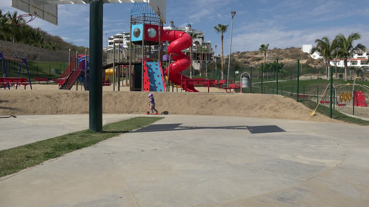 Helmeted child on a scooter near a colorful playground.