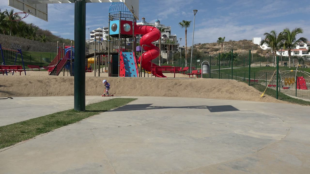 The child on the scooter moves along the lower left of the Los Cabos park frame, while the red playground slide and fenced sandy play area stay centered in bright sun.