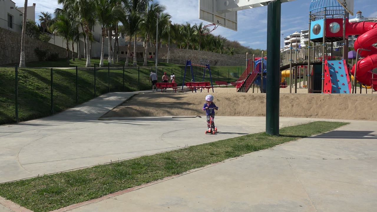 A young girl in a helmet rides a red scooter on a concrete path in Los Cabos, Mexico, with a playground, benches, grass, palms, and buildings behind her.