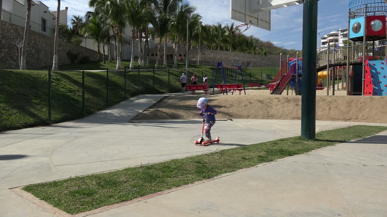 The helmeted girl continues across the concrete path in Los Cabos, Mexico, with the red playground structure and palm-lined slope farther back.