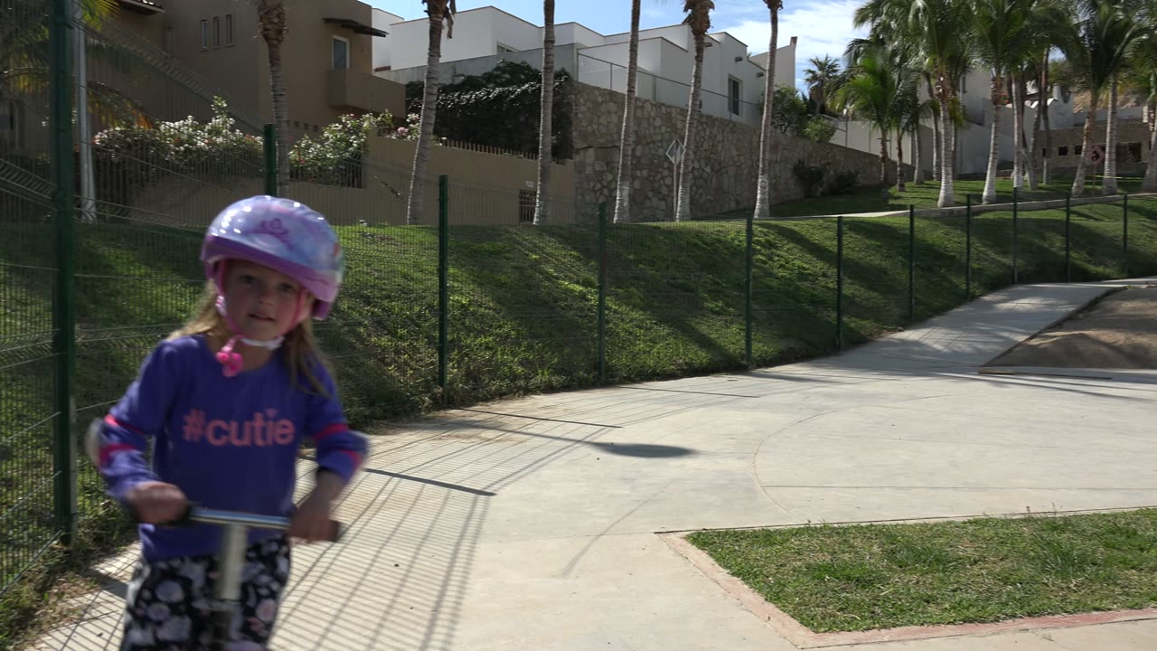 Young girl in a pink helmet and purple-blue shirt stands with a scooter near a paved path in Los Cabos, Mexico, with palms and hillside buildings behind her.
