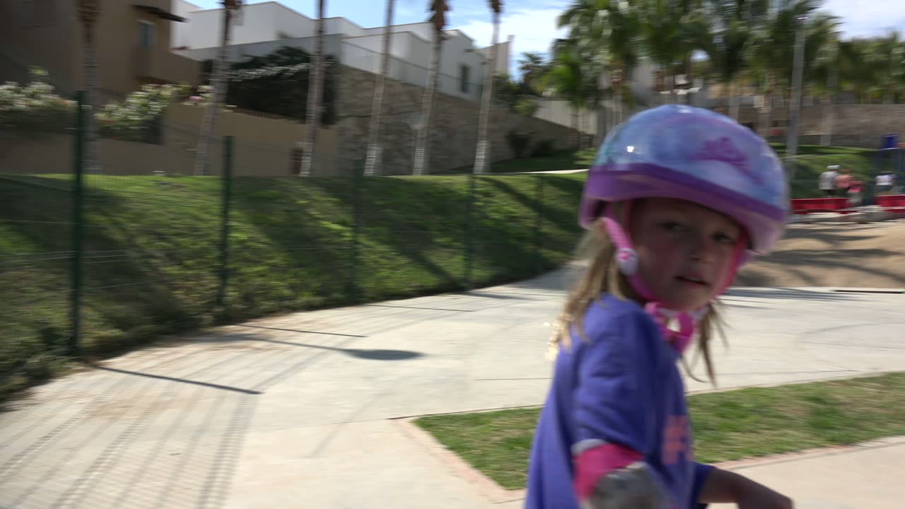 The girl rides close past the ground-level camera in Los Cabos, Mexico, with a concrete walkway, grass, palms, and red playground equipment in the background.