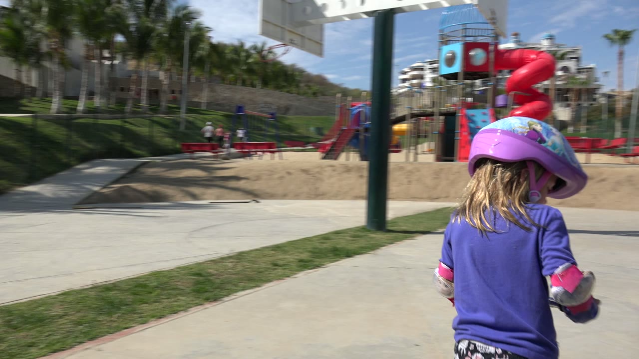Girl seen from behind riding scooter toward a playground.