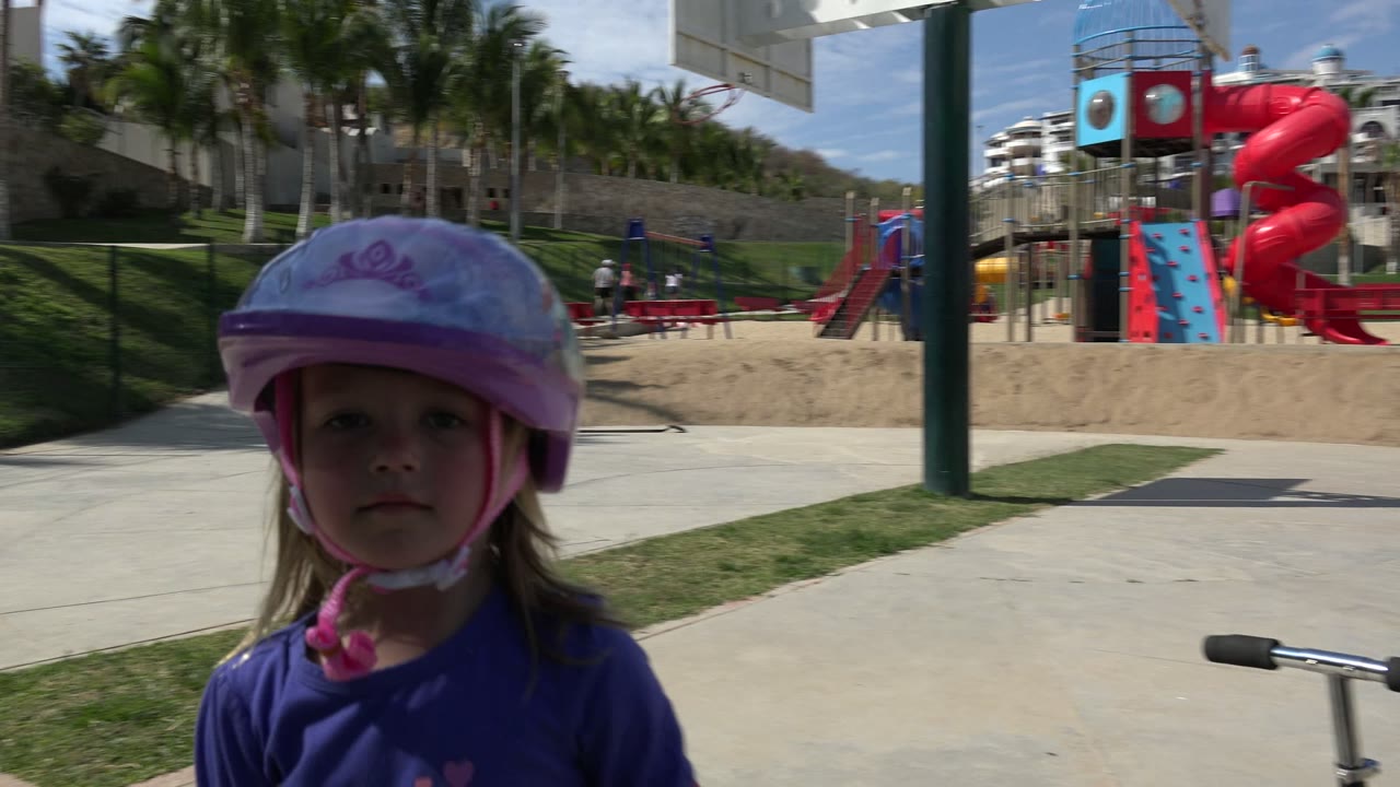 Young girl in a purple helmet near a paved playground path.