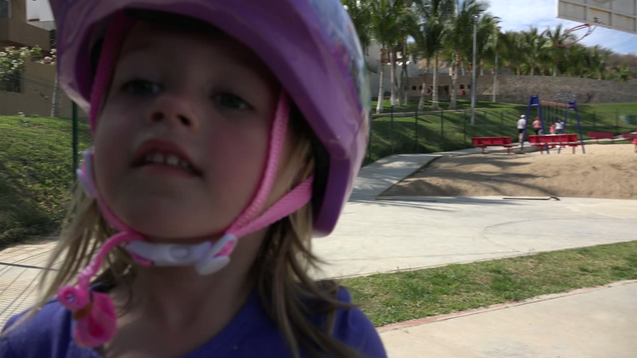 Close foreground view of the child in a purple helmet in Los Cabos, Mexico, with part of a scooter handlebar visible and the playground receding behind her.