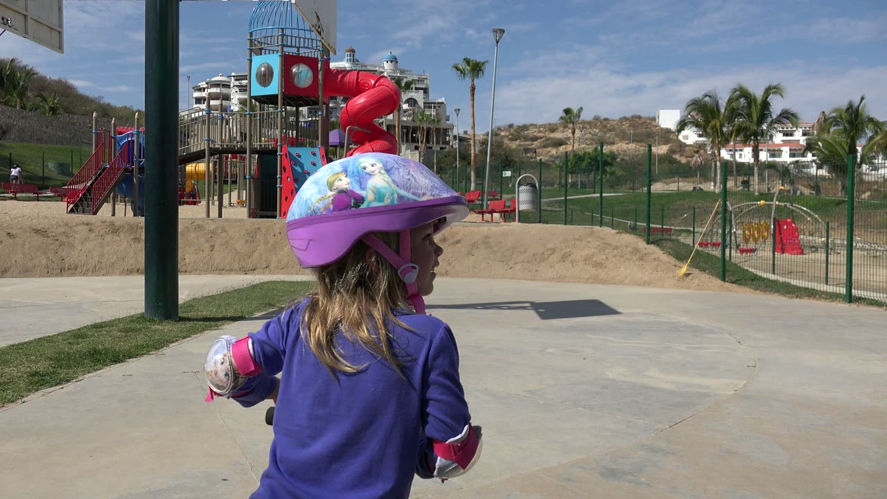 The child shifts toward the center foreground in the Los Cabos park, with the colorful helmet, purple shirt, red playground slide, fenced sand area, palms, and hillside buildings visible.