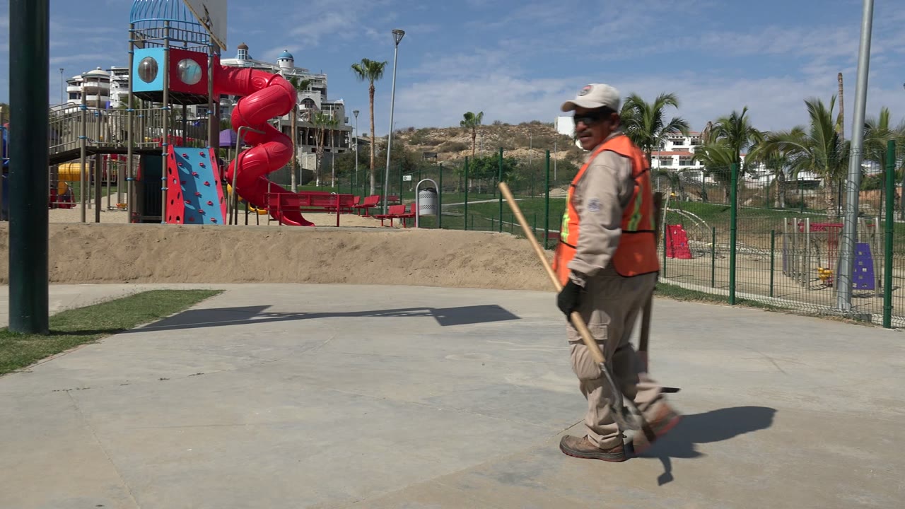 Groundskeeper walking past a red playground slide in a sunlit park.