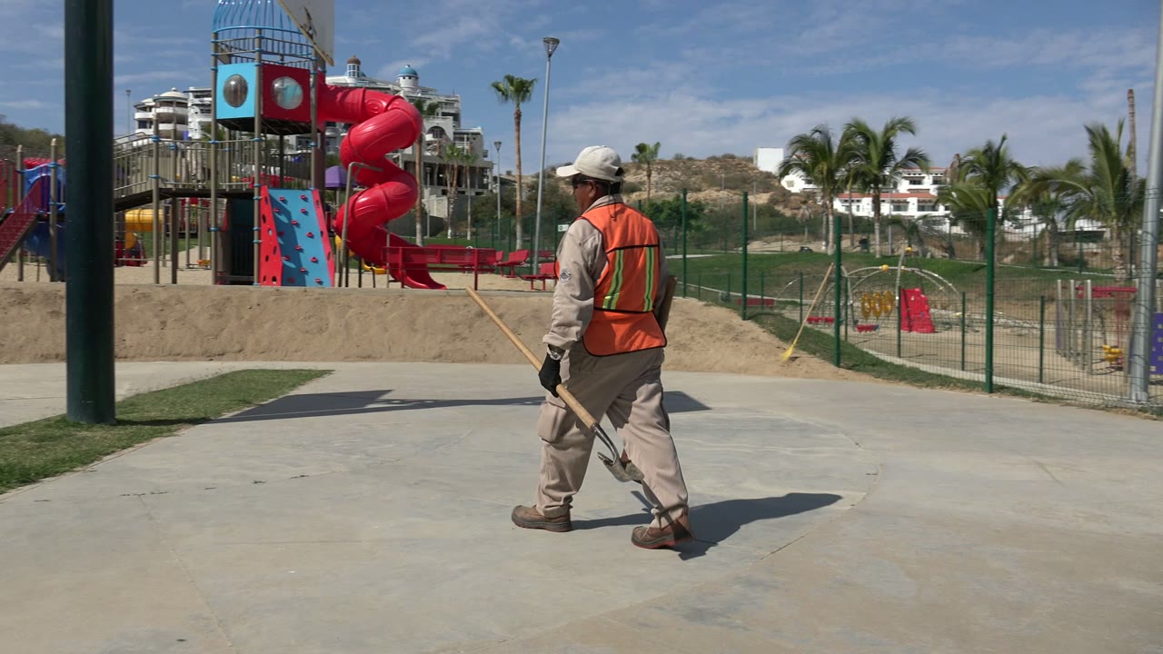 The worker continues across the concrete walkway in Los Cabos, holding tools near a fenced playground with a red slide, benches, palms, and dry hillside in view.