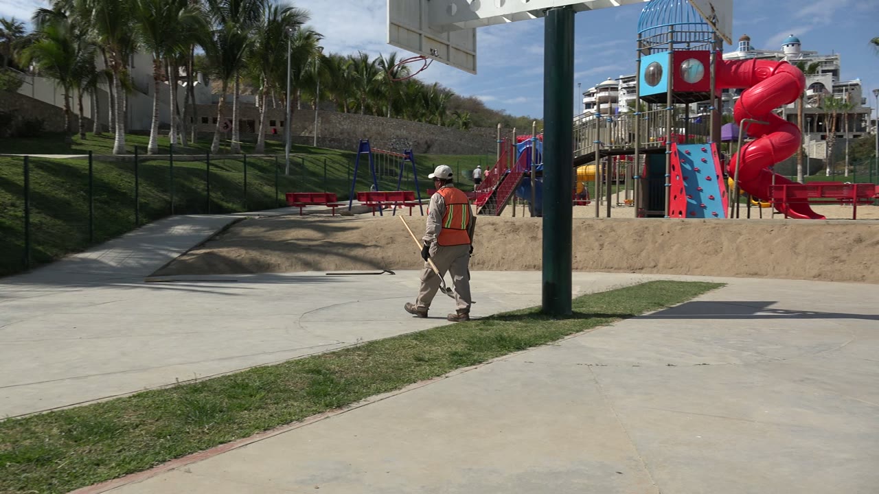 A worker in a safety vest crosses a paved area in Los Cabos, Mexico, with grass, palm trees, fencing, benches, and a colorful playground behind him.