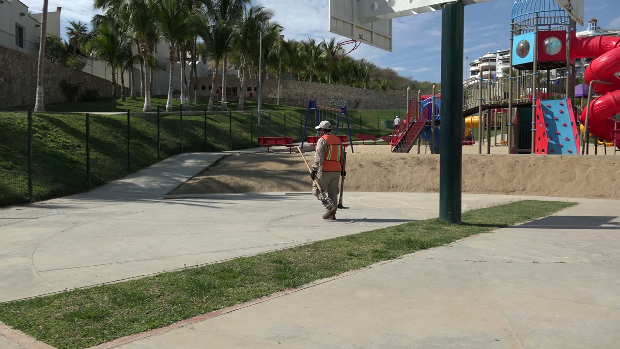 The worker moves farther across the concrete in Los Cabos, Mexico, with a red spiral slide, climbing panels, palms, and a green post visible around the playground.