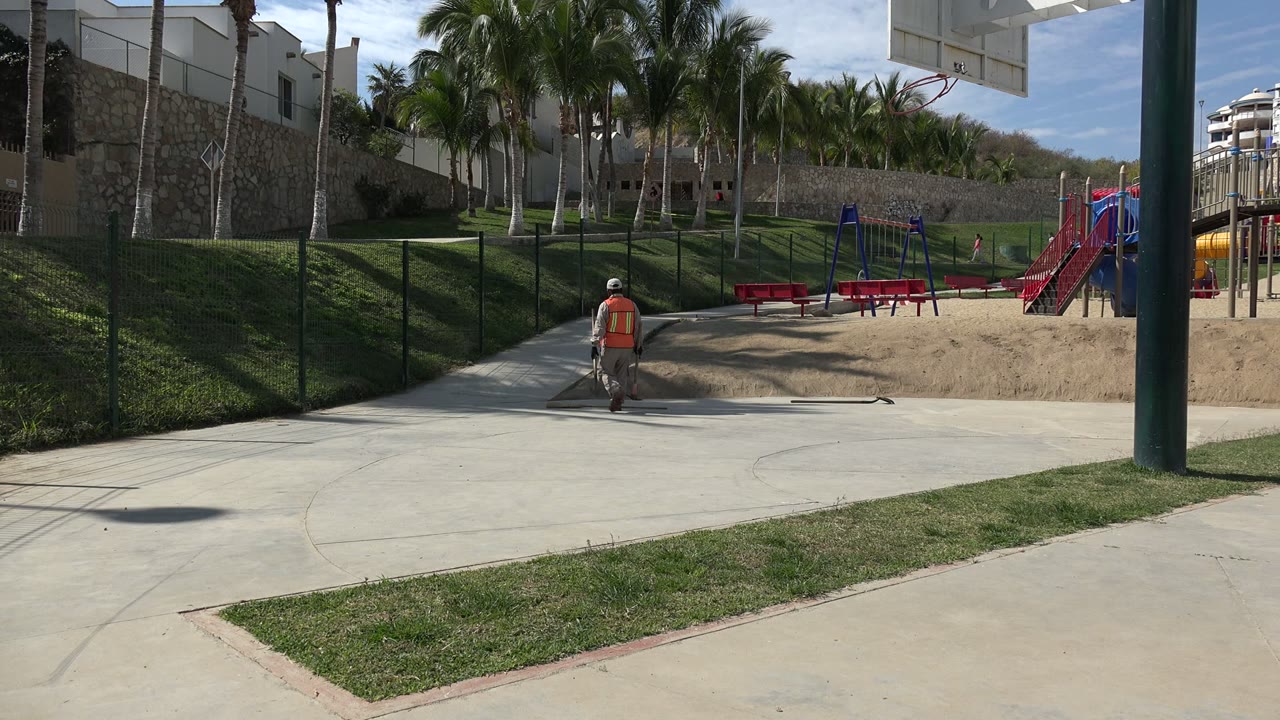 At 0:00 in Los Cabos, Mexico, a worker in an orange vest walks along a paved path beside sand, grass, fencing, palm trees, benches, and playground equipment.