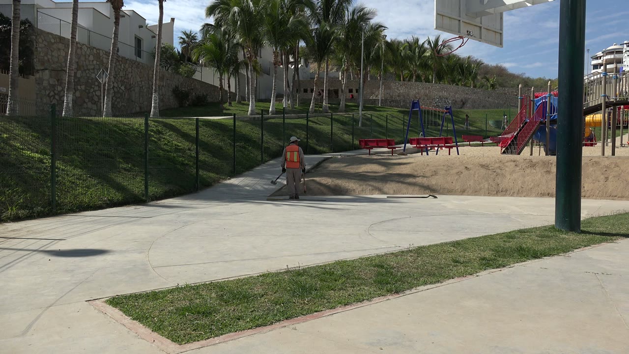 At 0:01 in Los Cabos, Mexico, the worker stands closer to a long material lying on the pavement while the sand play area, red benches, palms, fence, and buildings remain visible.