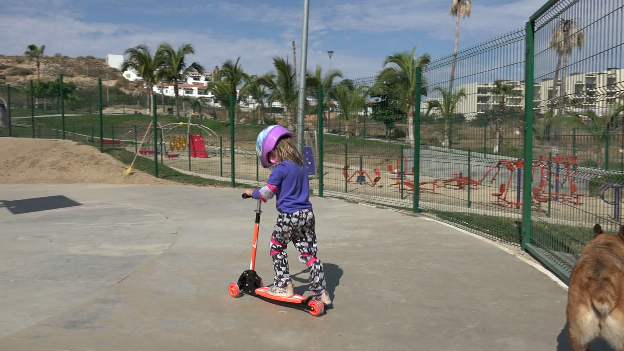 The helmeted girl continues riding the orange scooter across the concrete in Los Cabos, Mexico, with the brown dog partly visible near the right foreground fence.