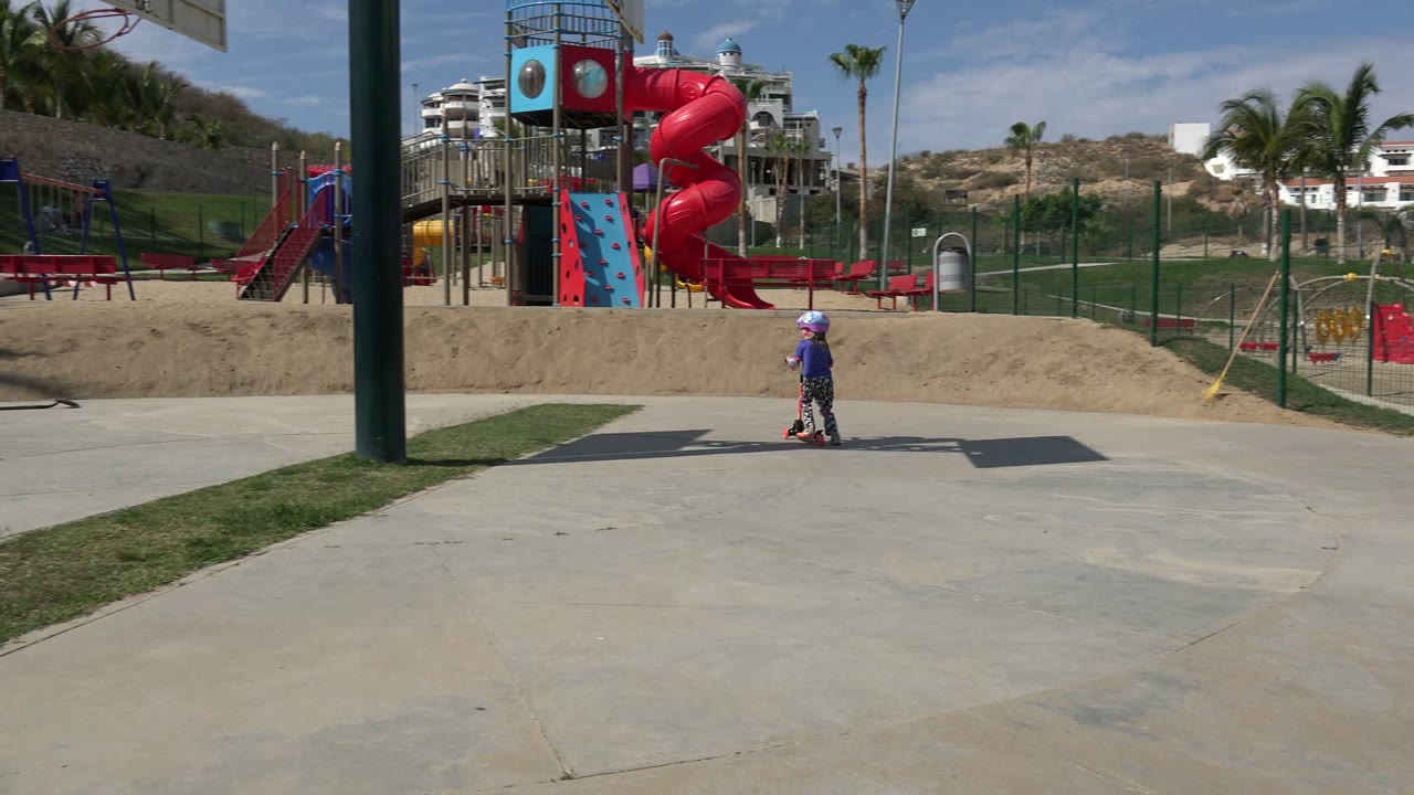 In Los Cabos, Mexico, a child in a helmet rides across a paved walkway near a fenced playground with a red spiral slide, benches, palms, and hillside landscaping.