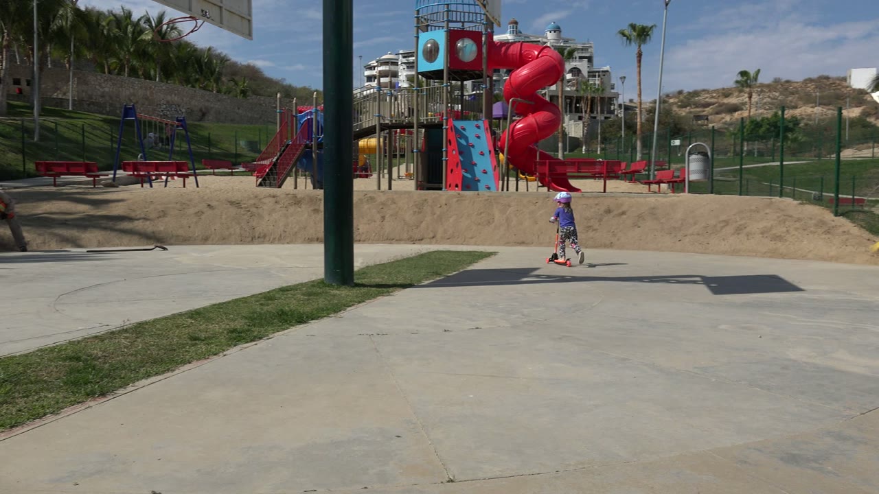 In Los Cabos, Mexico, the child rides farther along the paved path as an adult bends near the walkway by the fenced playground and red slide.