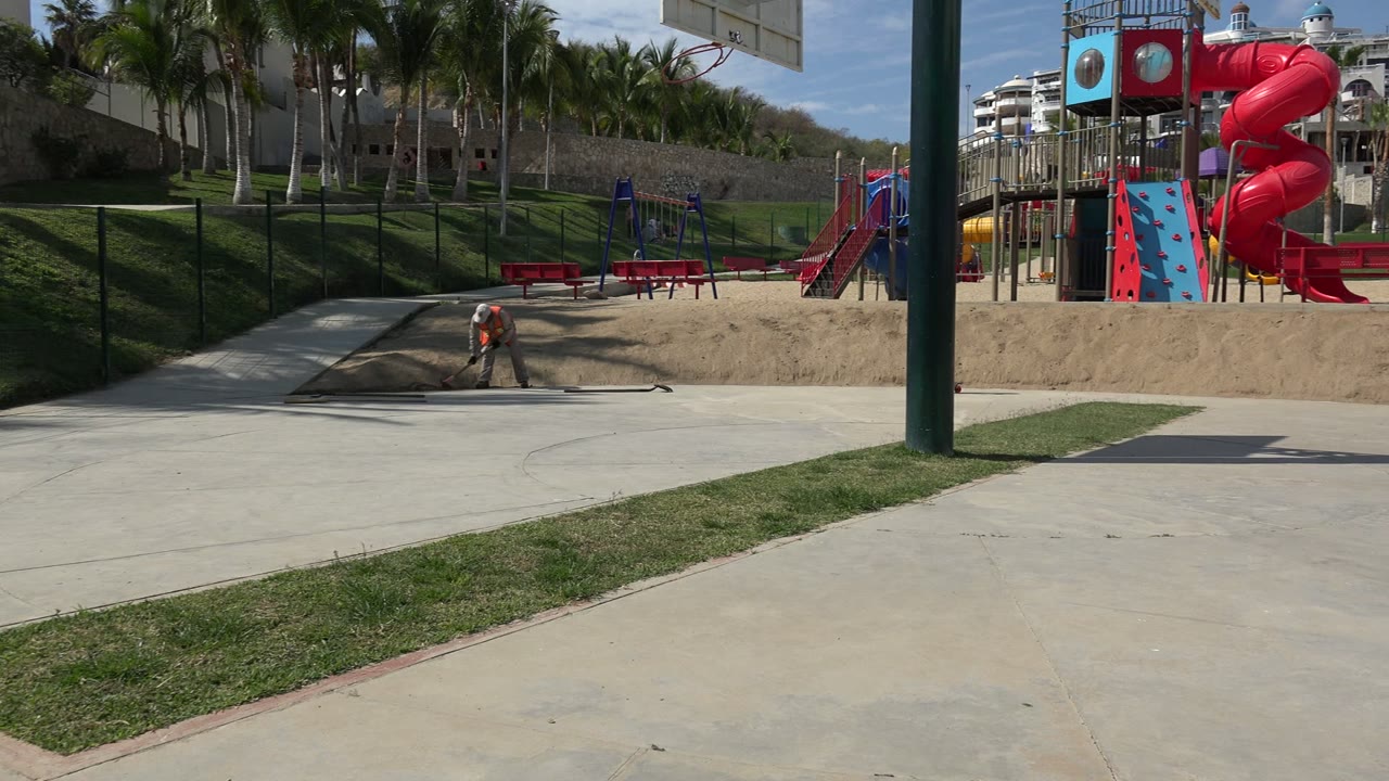 Wide playground path with an adult near sand and red slides in the background.