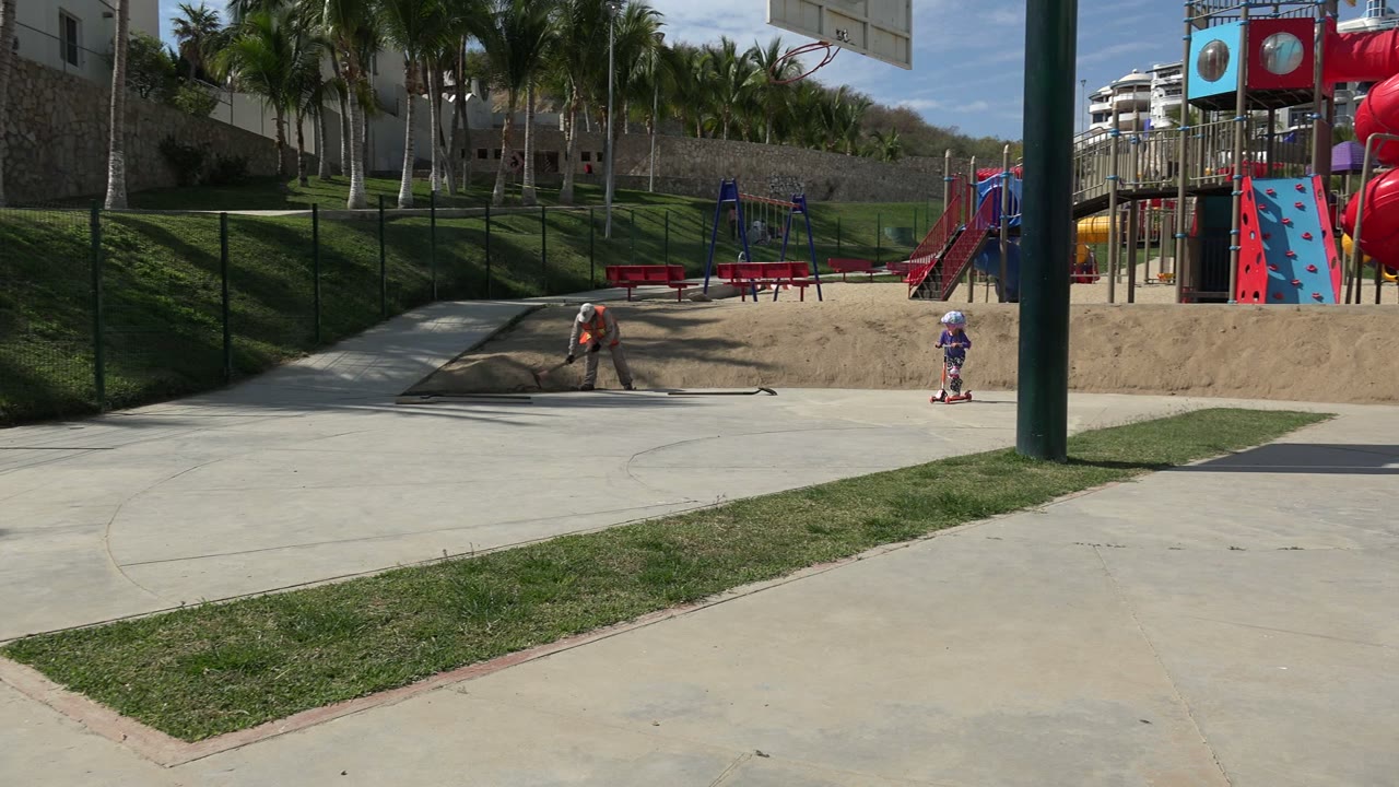 A helmeted child on a small scooter enters the Los Cabos playground frame from the right, while an adult remains near the sandy play area and red slides behind.