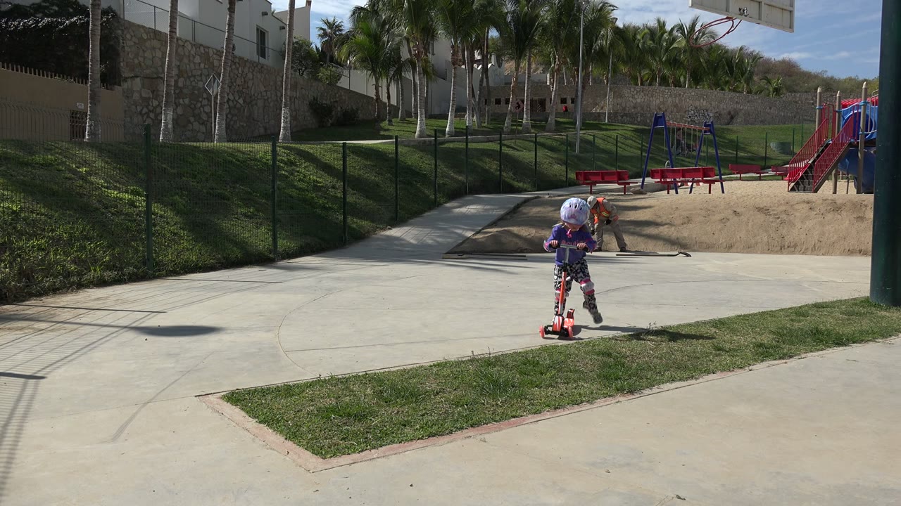 Helmeted child on a red scooter on a paved walkway near a playground.