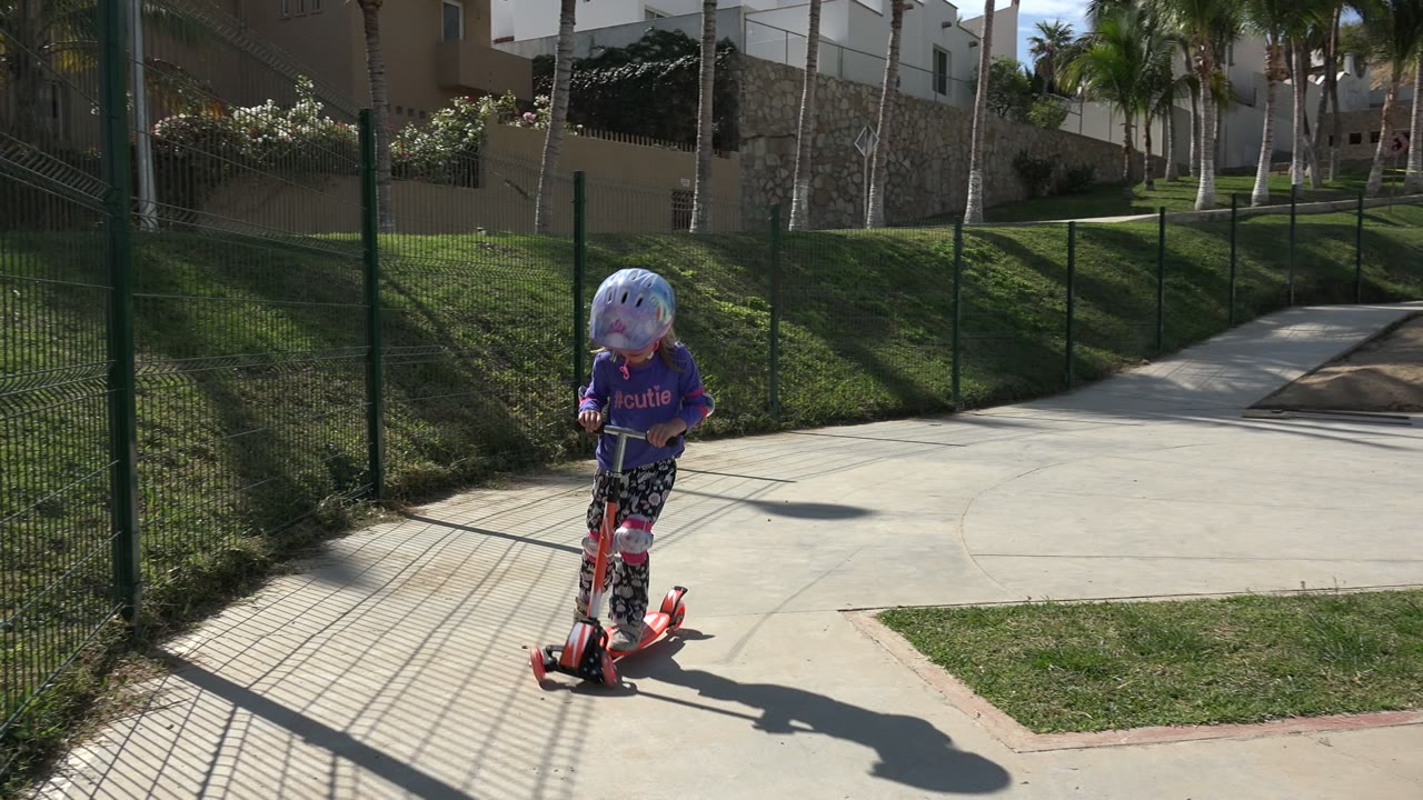 A young girl in a purple shirt, pink helmet, and protective pads rides a small orange scooter along a paved play area in Los Cabos, Mexico, with grass, fencing, and buildings behind her.