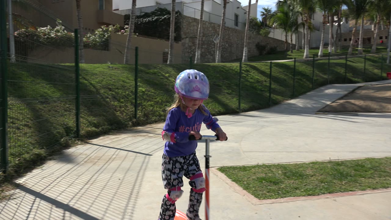 The helmeted girl rides closer to the camera in Los Cabos, Mexico, with the scooter handlebars in front of her and a fenced grassy slope, sidewalk, palms, and buildings in the background.
