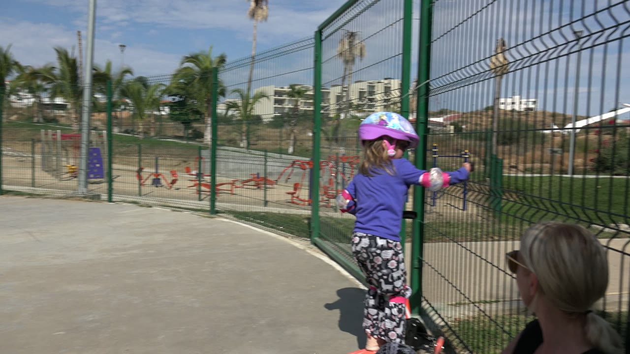In Los Cabos, Mexico, a helmeted young girl stands beside a small skating bike near a green metal fence while an adult woman sits in the foreground.