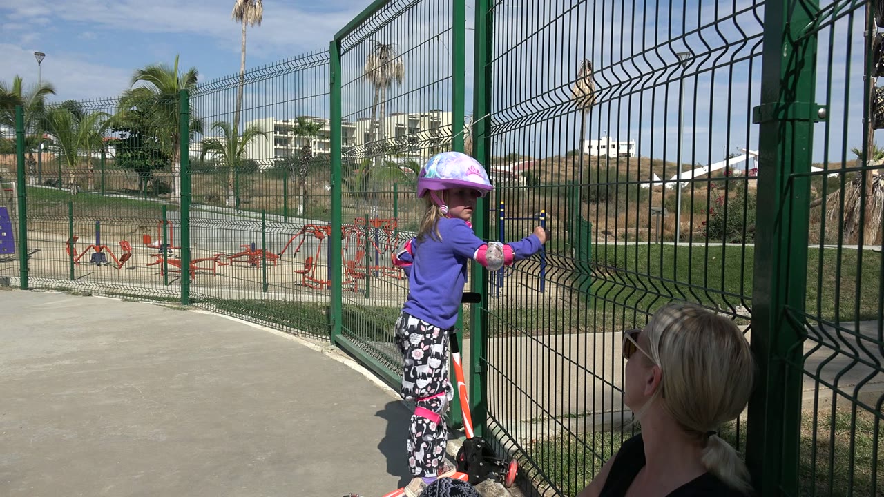 In Los Cabos, Mexico, the girl turns toward the fence with one hand on the small skating bike, while the seated adult remains partly visible in the lower right.