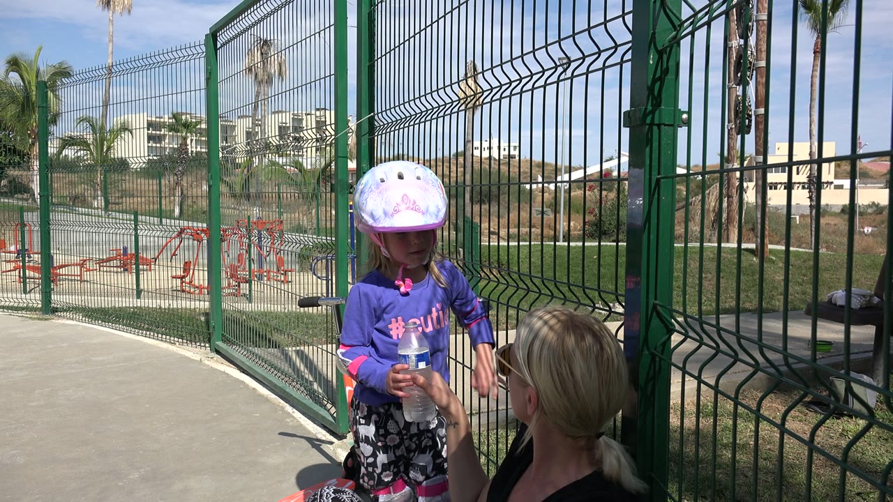 Child in helmet holding a water bottle beside a seated adult at a fenced skate park.