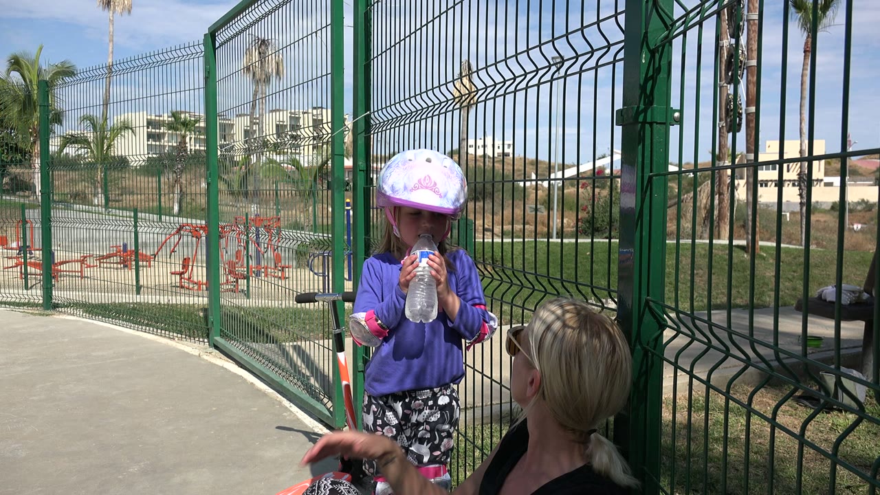 At 0:01 in Los Cabos, Mexico, the child lifts the clear bottle to drink as the adult remains seated at the lower right beside the green fence.