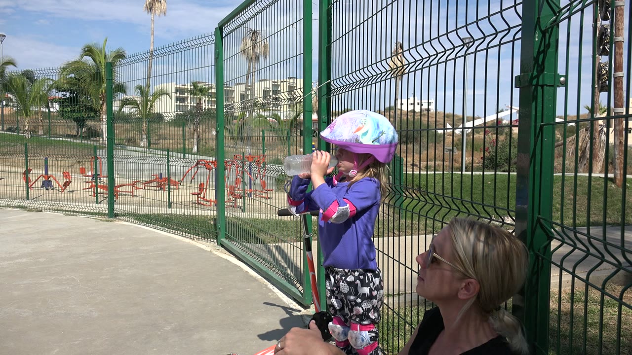 In Los Cabos, Mexico, a young child wearing a colorful helmet drinks from a clear bottle while a seated adult woman is partly visible beside a green park fence.