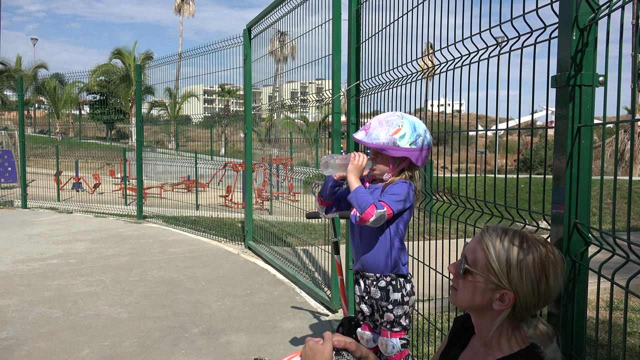In Los Cabos, Mexico, the helmeted child remains at the fence drinking water, with the adult woman lower in frame and palm trees and playground equipment behind the fence.