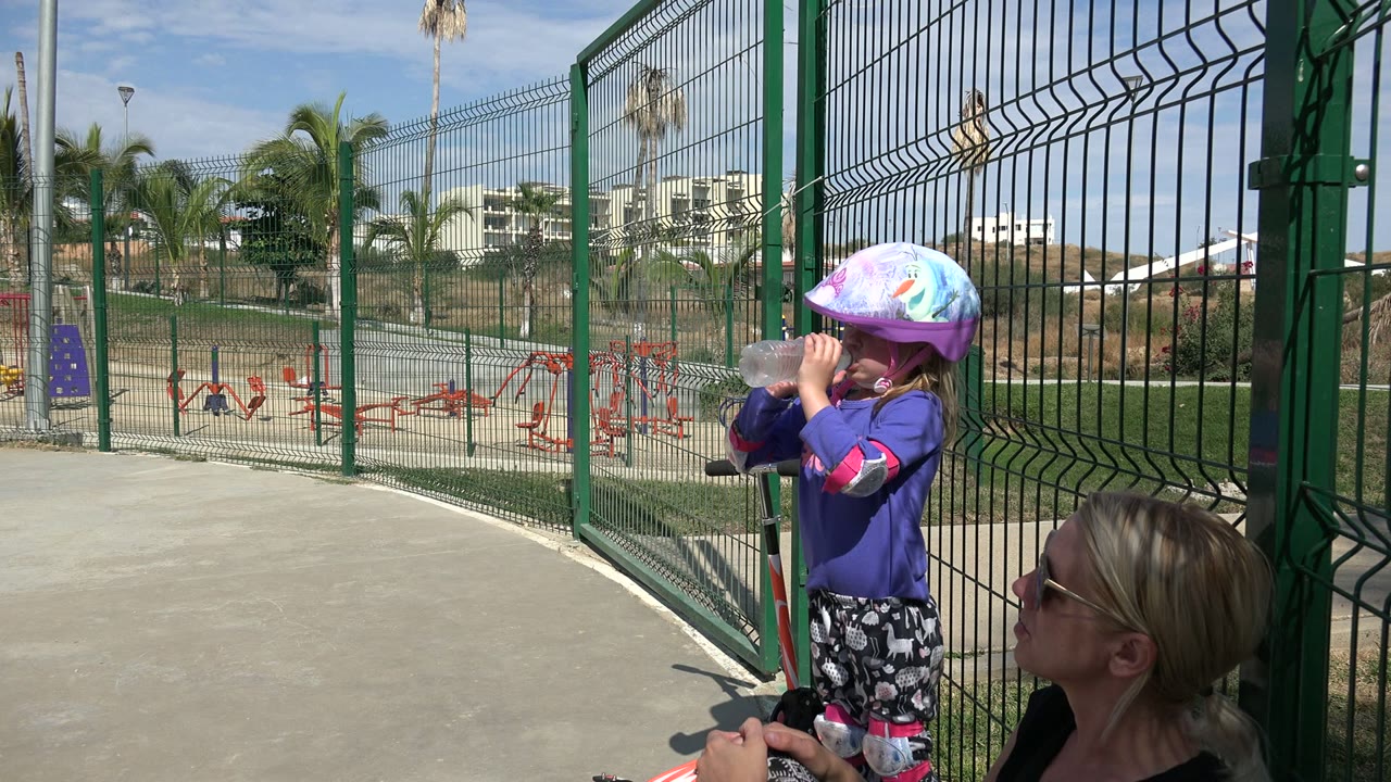 Child in protective gear drinks water near a tall green fence and playground.