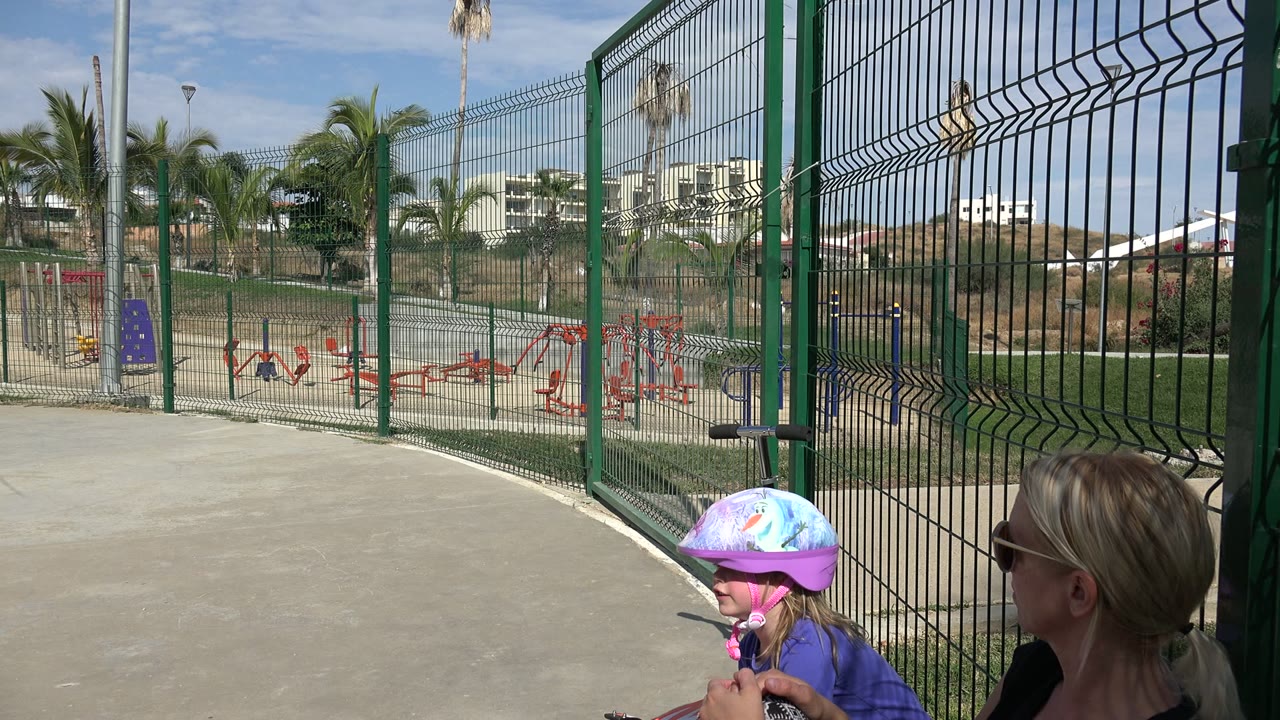 Young girl in a helmet beside her mother near a fenced playground.