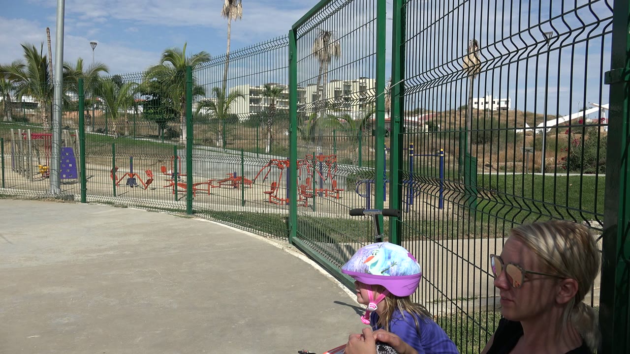 At 0:01 in Los Cabos, the helmeted girl remains near the lower right while her mother sits beside her, with the fence bars and red playground structures clearly visible.