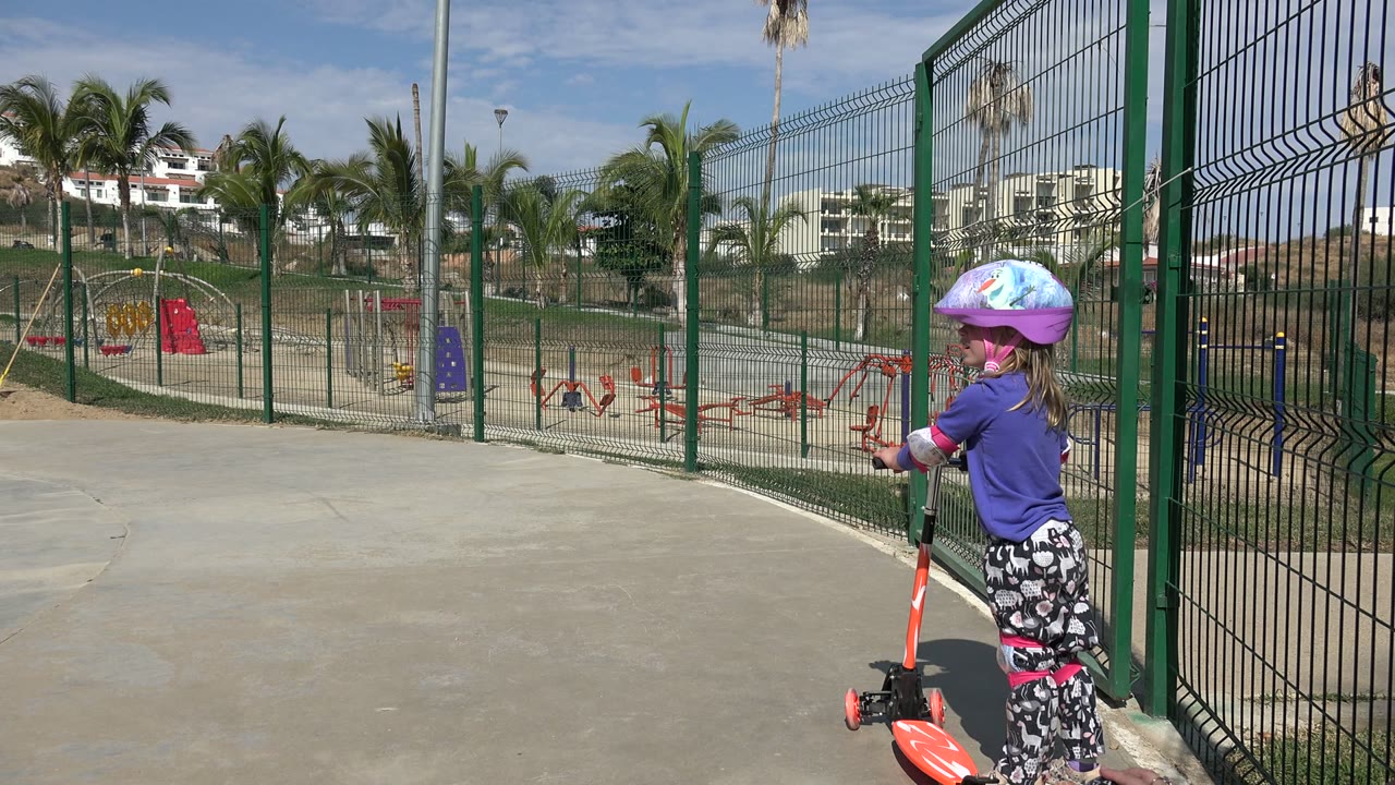 A young girl in a colorful helmet holds an orange scooter on sunlit concrete beside a green metal fence in Los Cabos, Mexico, with playground equipment and palm trees behind her.