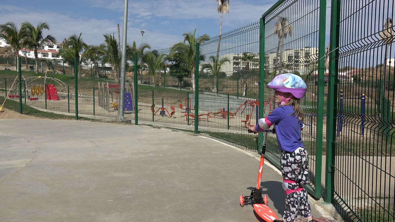 The helmeted girl remains beside the green fence with one hand on the scooter handlebar, while the park playground, palm trees, and hillside buildings stay visible in the background in Los Cabos, Mexico.