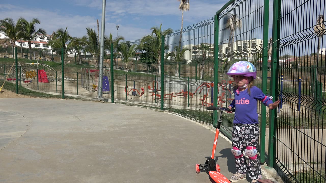 Girl facing the camera with scooter beside fenced playground.