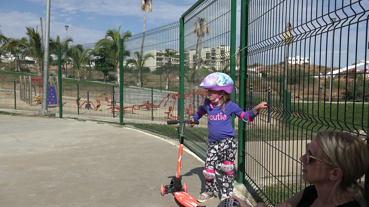 Young girl with helmet standing beside an orange scooter near a green fence.