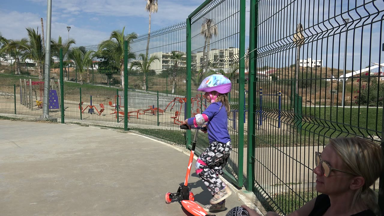 In Los Cabos, the helmeted child begins turning the orange scooter along the concrete path beside the green fence, with the seated adult woman still visible at frame edge.